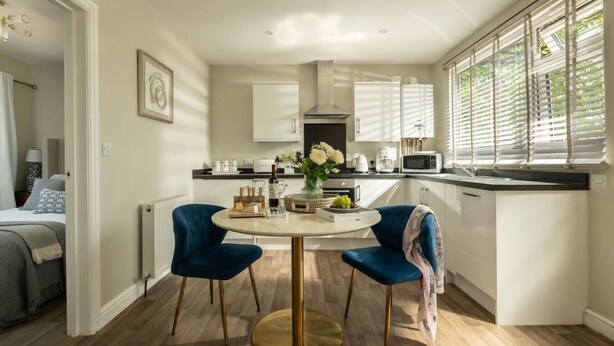 A modern kitchen with white cabinets, a dining table with two blue chairs, and a view of the outdoors through the window.