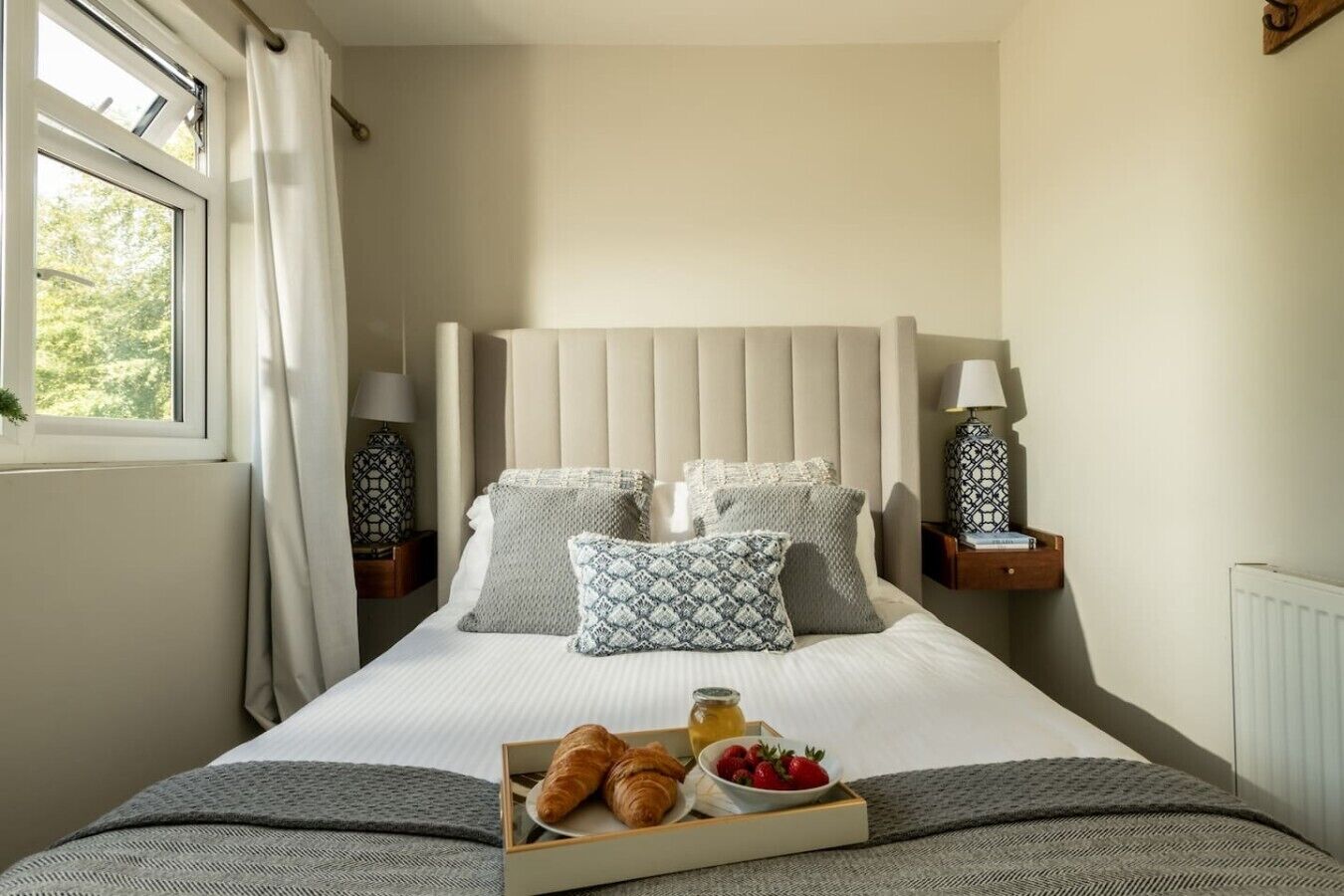 A neatly made bed with a tray of croissants and strawberries, two bedside lamps, and a window with curtains.