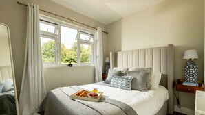 A neatly made bed with pillows and a tray of breakfast items, a bedside table with a lamp, and a window with curtains.