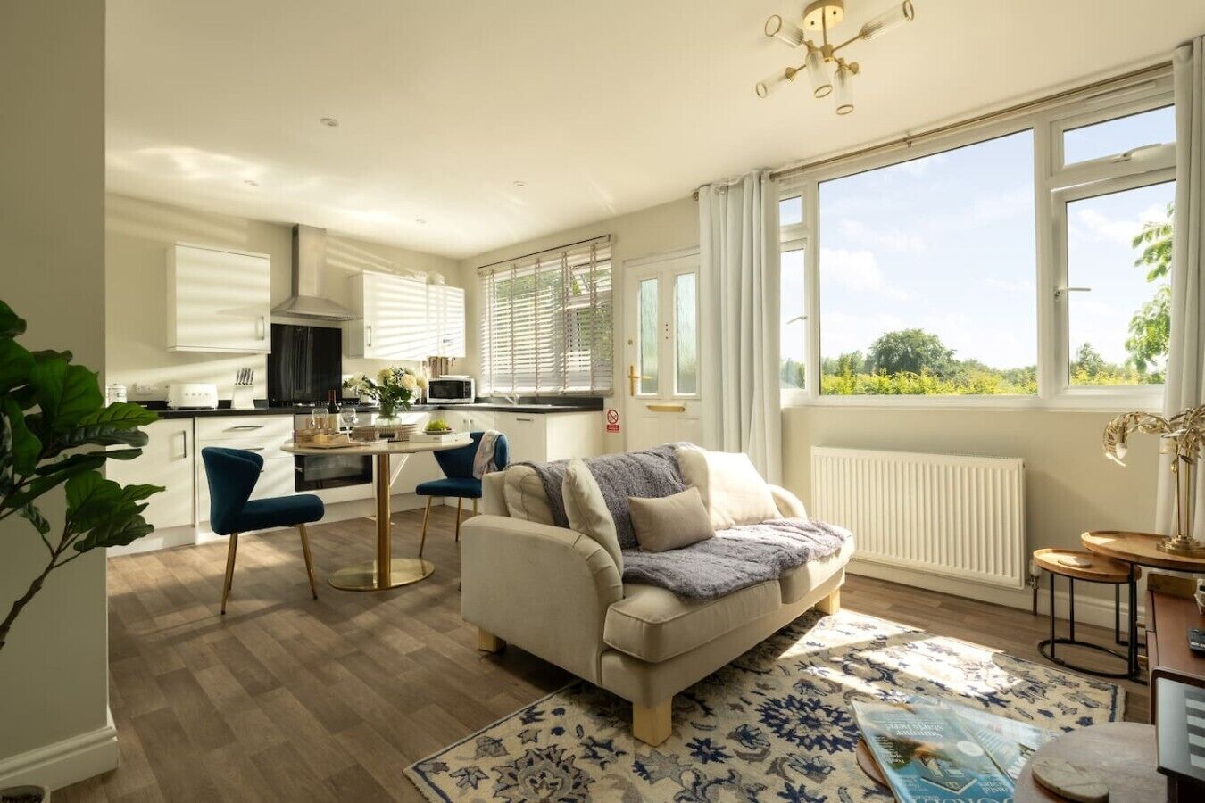 A modern kitchen with white cabinets, a dining area with a blue chair, and a large window offering a view of greenery.