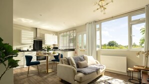 A modern kitchen with white cabinets, a dining area with a blue chair, and a large window offering a view of greenery.