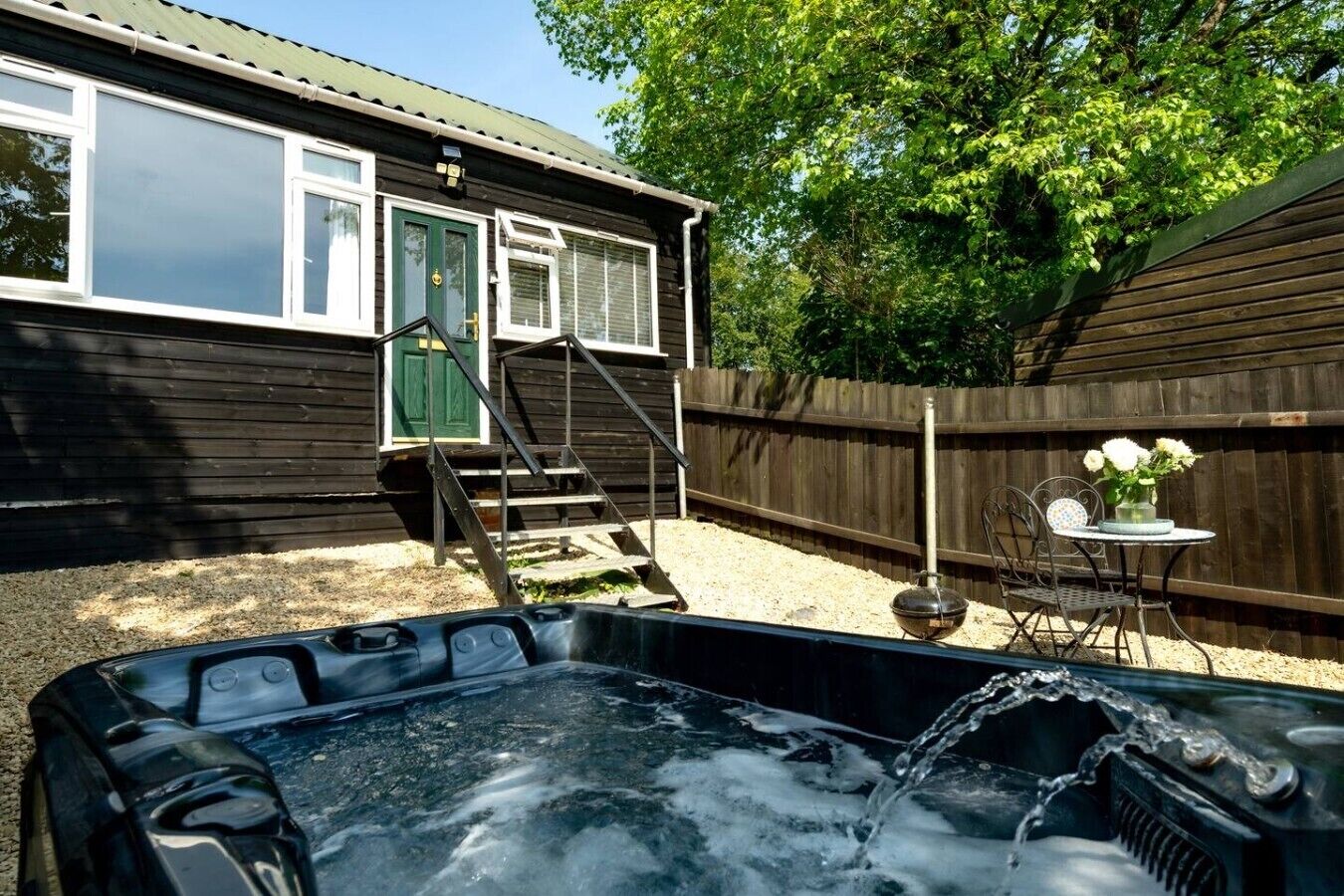 Ein Whirlpool mit fließendem Wasser, eine Holzdielen-Terrasse, ein Metallstuhl, ein Tisch mit Blumen und ein Haus mit grüner Tür und Fenstern.