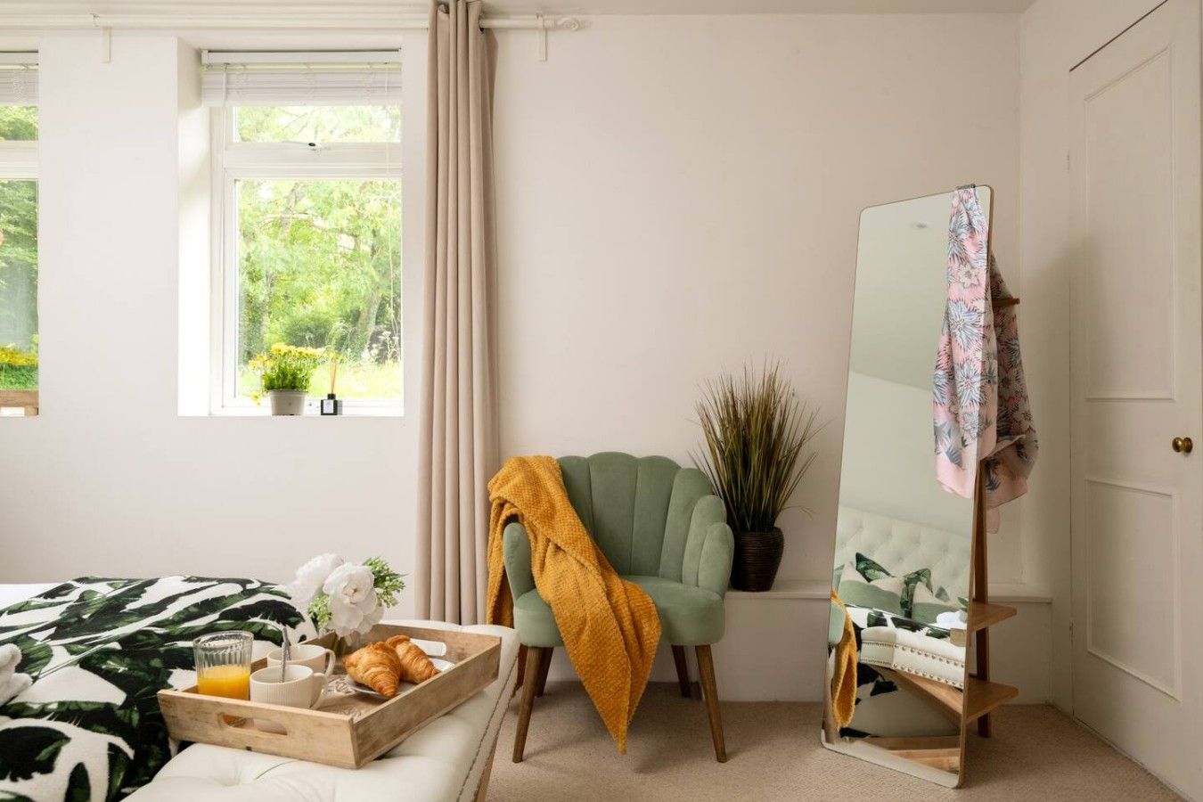 A bedroom with a bed, a bedside table with a tray of croissants and orange juice, a green armchair with a yellow throw, a potted plant, and a mirror on a stand.