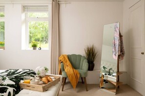 Un dormitorio con cama, una mesita de noche con una bandeja de cruasanes y zumo de naranja, un sillón verde con una manta amarilla, una planta en maceta y un espejo sobre un soporte.