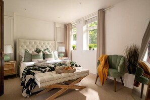 A bedroom with a tufted headboard, bedside tables with lamps, a green armchair, a window with curtains, and a small plant.