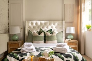 A neatly made bed with a tufted headboard, two bedside tables with lamps, and a window with a view of greenery.