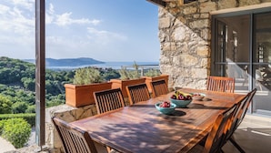 Une table en bois, entourée de chaises, avec un saladier rempli de fruits, le tout placé devant un mur de pierre offrant une vue sur la mer et les montagnes.