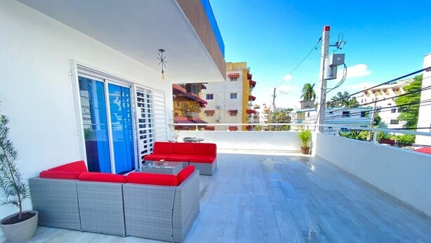 A balcony with grey wicker furniture and red cushions, a potted plant, and a view of buildings under a clear sky.