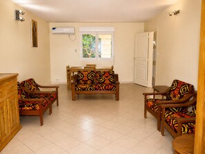 Een kamer met een zithoek, een eettafel en stoelen, een raam met uitzicht op bomen, en een airconditioning.