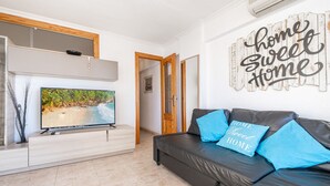 A living room with a TV, a black leather sofa with blue cushions, a wall decoration saying "home sweet home", and a door leading to another room.