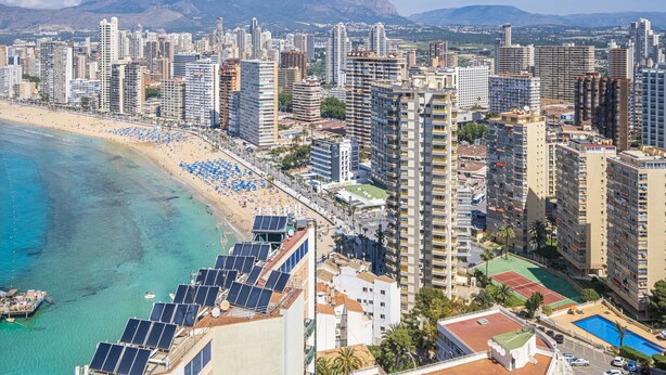 A coastal cityscape with high-rise buildings, a beach with blue umbrellas, and a mountain in the background.