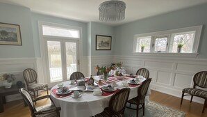 A dining room with a round table set for six, surrounded by six striped chairs. There is a chandelier above the table, and the room has large windows with white frames. - The Boardwalk Inn  (Summerside)