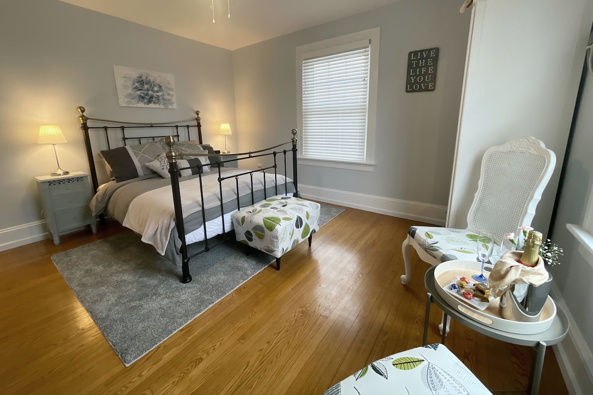Un dormitorio con cama con dosel, mesitas de noche con lámparas, una ventana con persianas, una silla con una mesa y una pared con una cita motivacional.