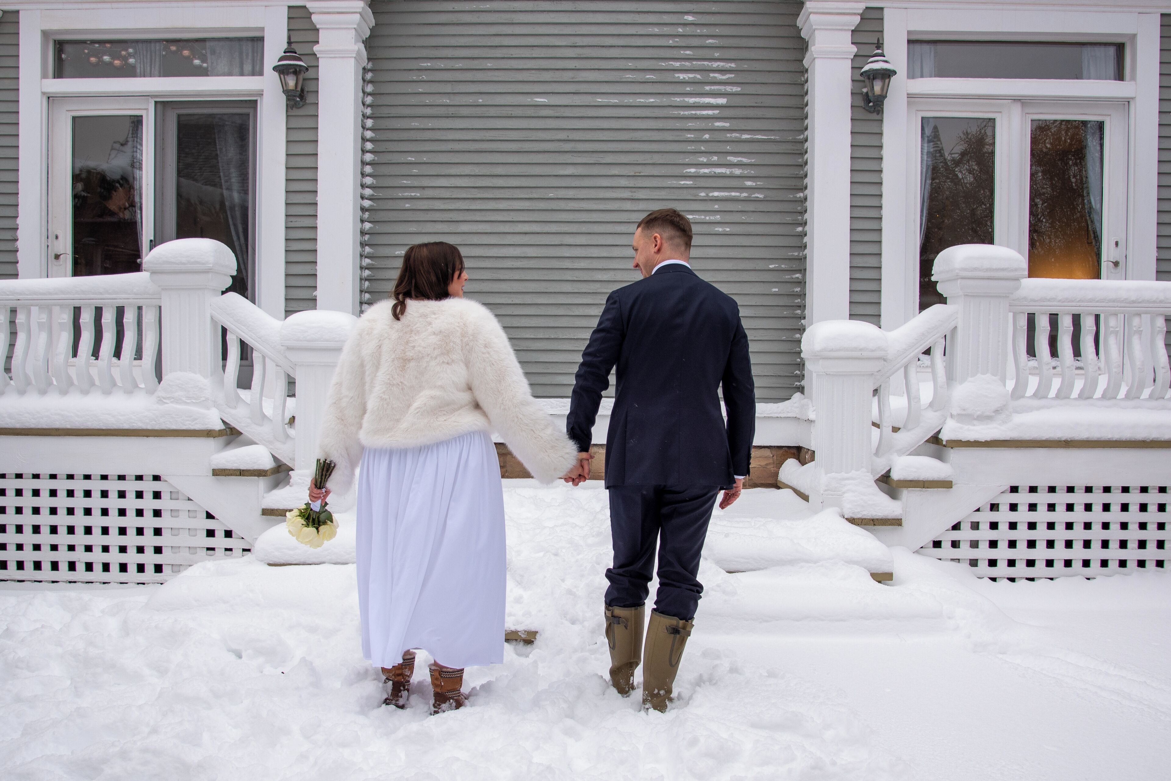 A couple holding hands in a snowy outdoor area with a white porch and steps.