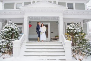 A white building with a porch, steps, and snow-covered trees.