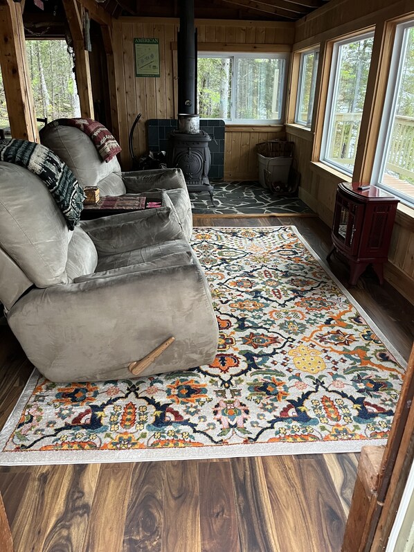 A cozy cabin interior with a fireplace, a sofa, a patterned rug, and a view of the forest through large windows.