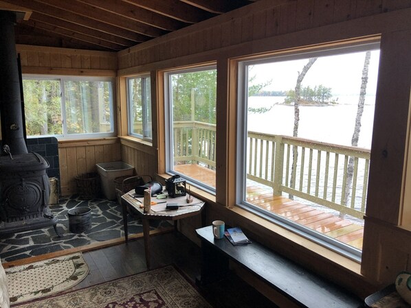 A cabin interior with a wood stove, a table with items, and large windows overlooking a wooden deck and lake.