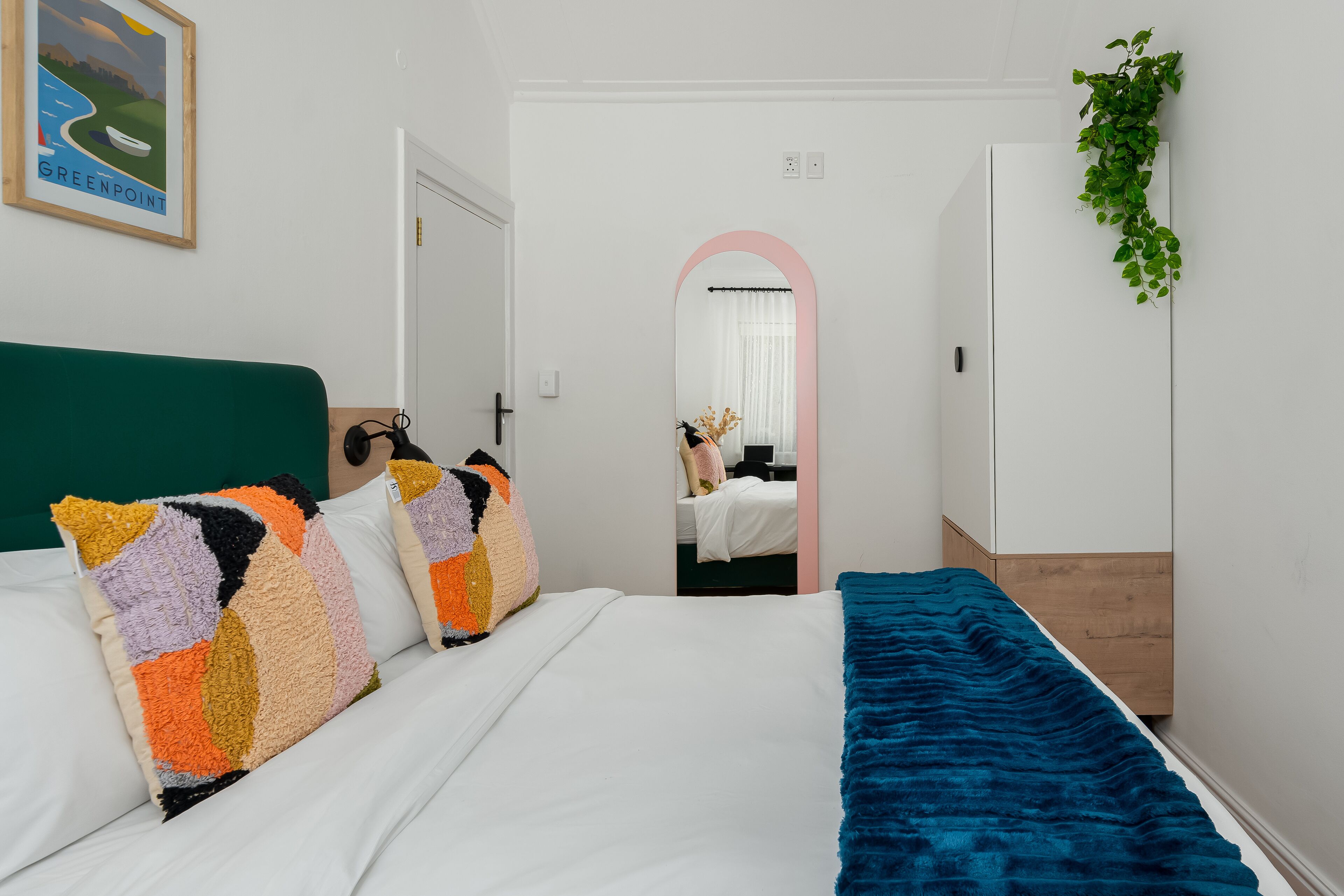 A bedroom with a bed, colorful pillows, a blue blanket, a mirror, and a green plant.