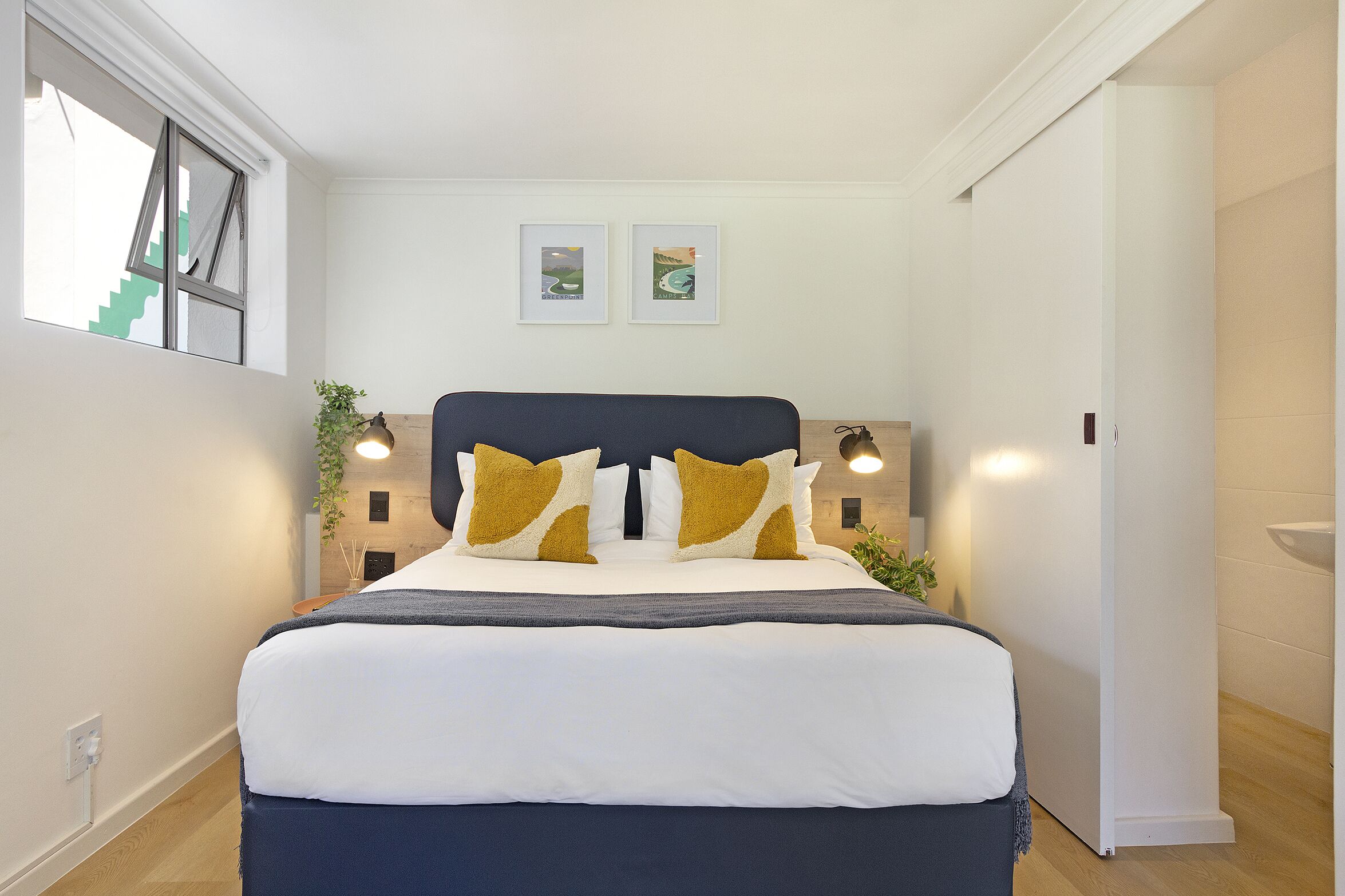 A modern bedroom with a large bed, two framed pictures on the wall, and a window with a view of stairs.