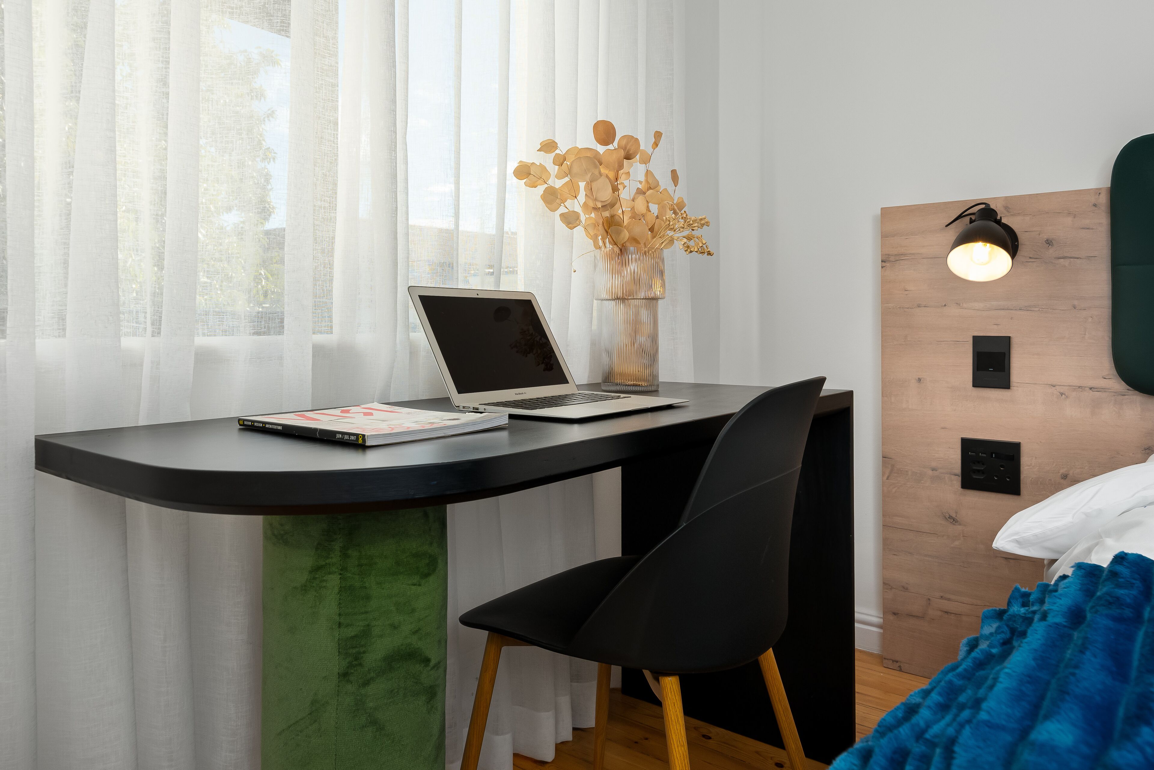 A modern bedroom with a curved desk, a laptop, a book, a vase with dried flowers, a black chair, a bed with a blue blanket, and a wall-mounted light.