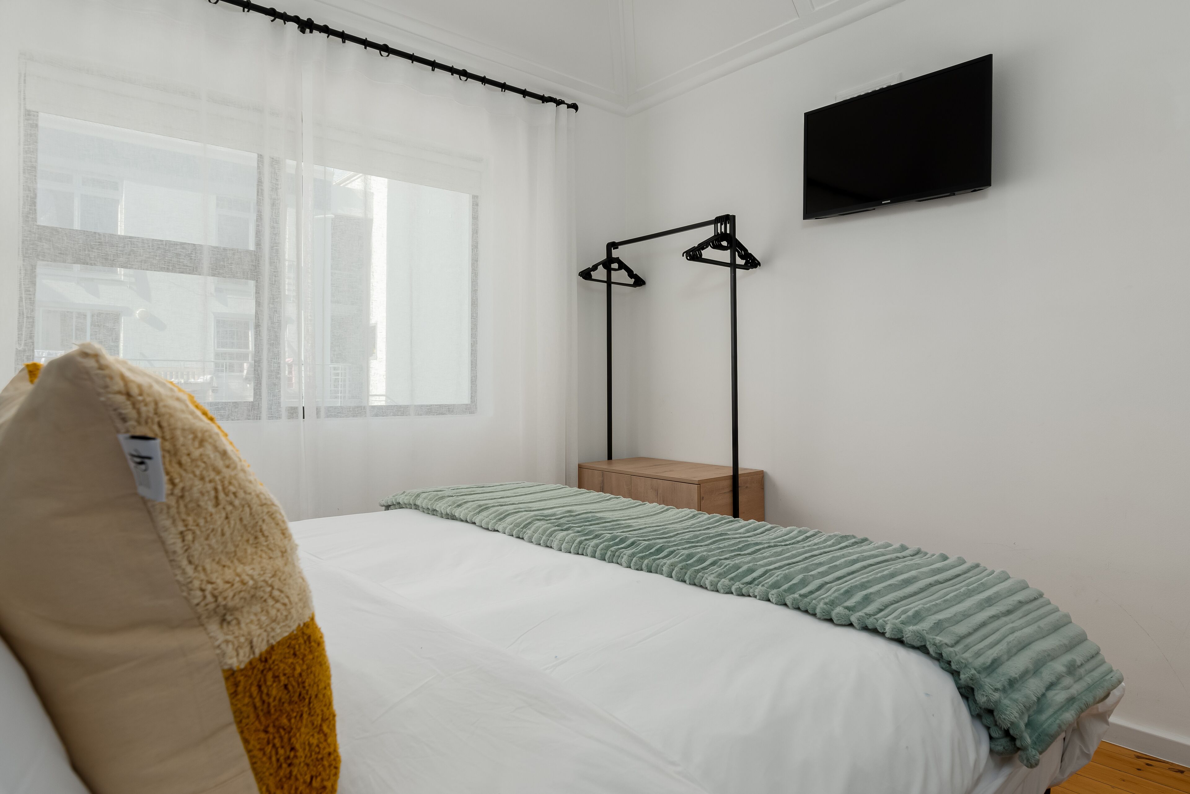 Una camera da letto con un letto, un comodino, una TV a parete e una finestra con tende leggere.
