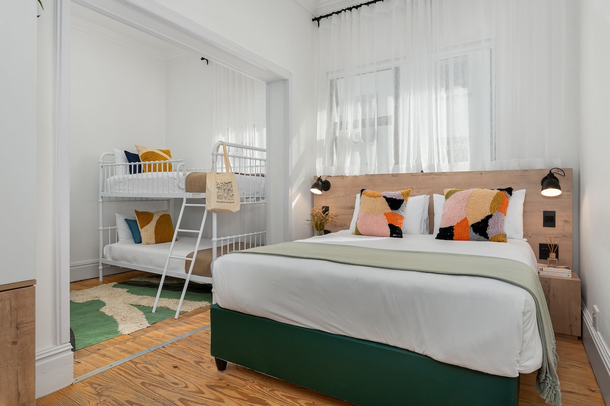 A bedroom with a bunk bed, a large bed with a wooden headboard, a green bed skirt, and a nightstand with a lamp.