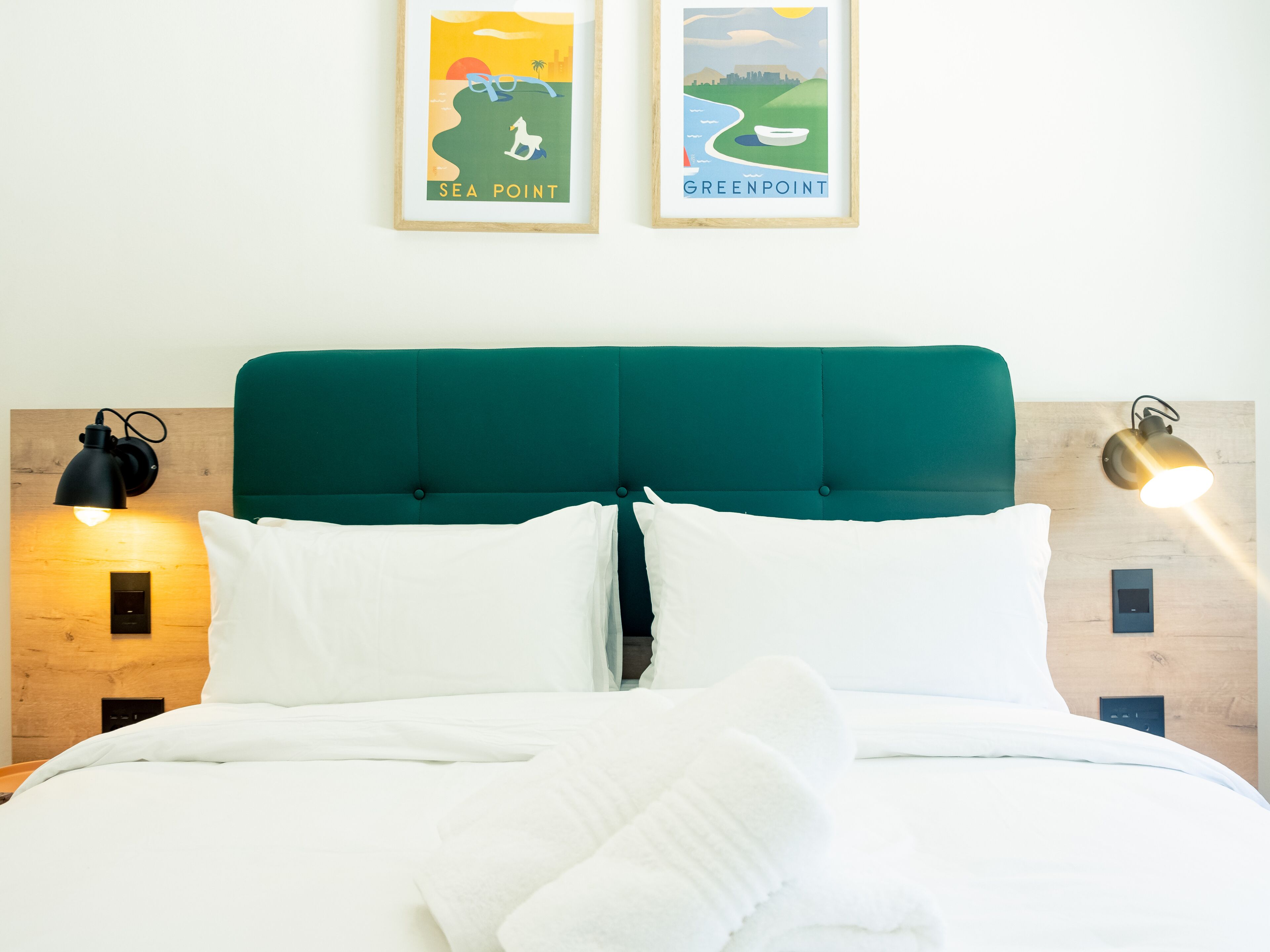 A neatly made bed with white linens and pillows, a teal upholstered headboard, and two wall-mounted reading lamps.