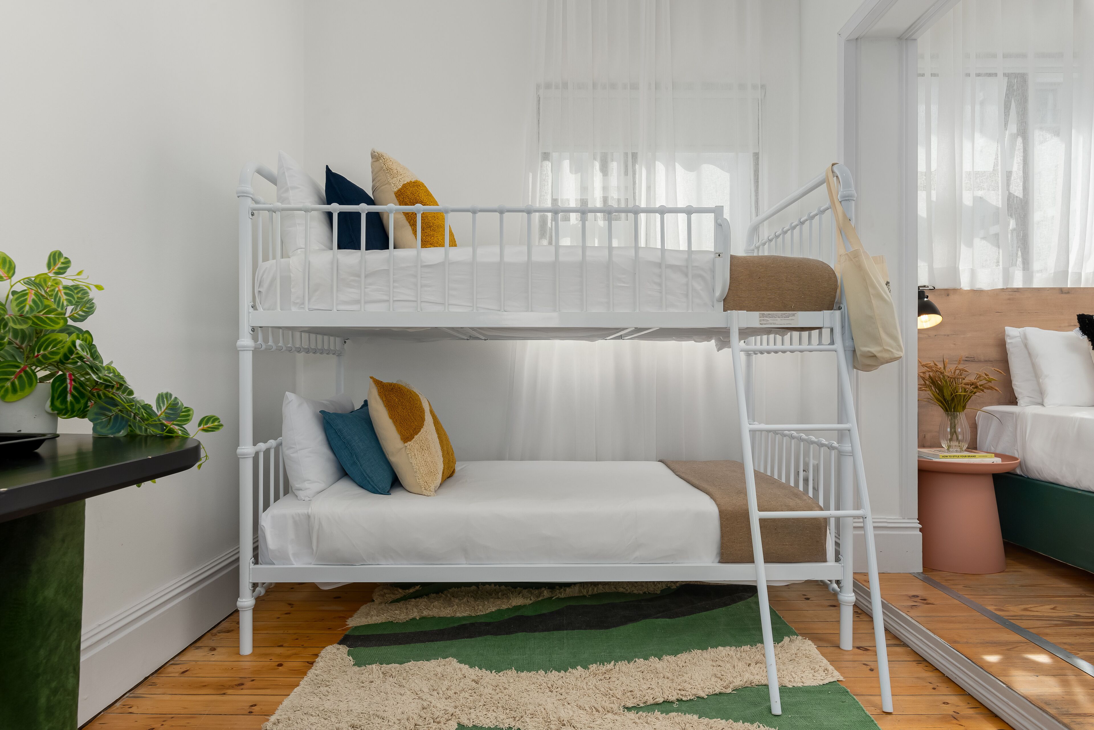 Un lit superposé avec une literie blanche, un tapis vert et beige, une plante en pot et une table de chevet.