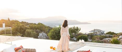 A woman in a long dress standing on a patio overlooking a scenic view of the sea and mountains.