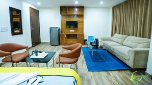 A hotel room with a bed, two chairs, a sofa, a TV mounted on a wooden cabinet, a refrigerator, and a coffee table. - Lime Tree Hotel Near 32nd Milestone (Gurugram)