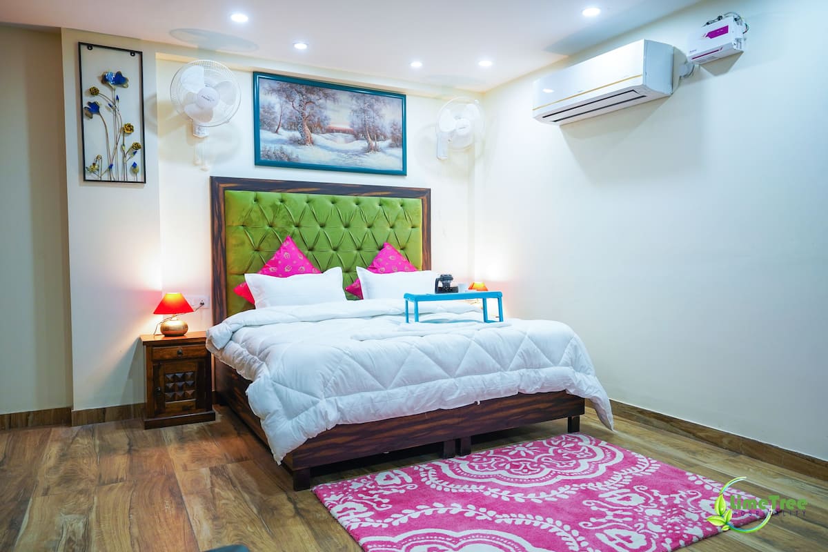 Un dormitorio con cabecera tapizada verde, ropa de cama blanca, alfombra rosa estampada, una mesita de noche con lámpara y un equipo de aire acondicionado.