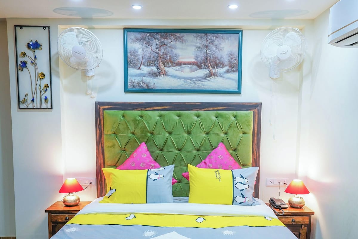 Un dormitorio con cabecera verde capitonada, cama con almohadas de colores, dos lámparas de noche, un cuadro enmarcado y ventiladores de pared.