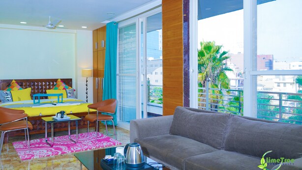 A modern hotel room with a sofa, dining table, chairs, and a balcony with a view of the city. - Lime Tree Hotel Near 32nd Milestone (Gurugram)