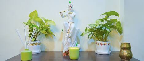 A statue of a deity, two potted plants, and a brass container on a wooden table against a plain wall.