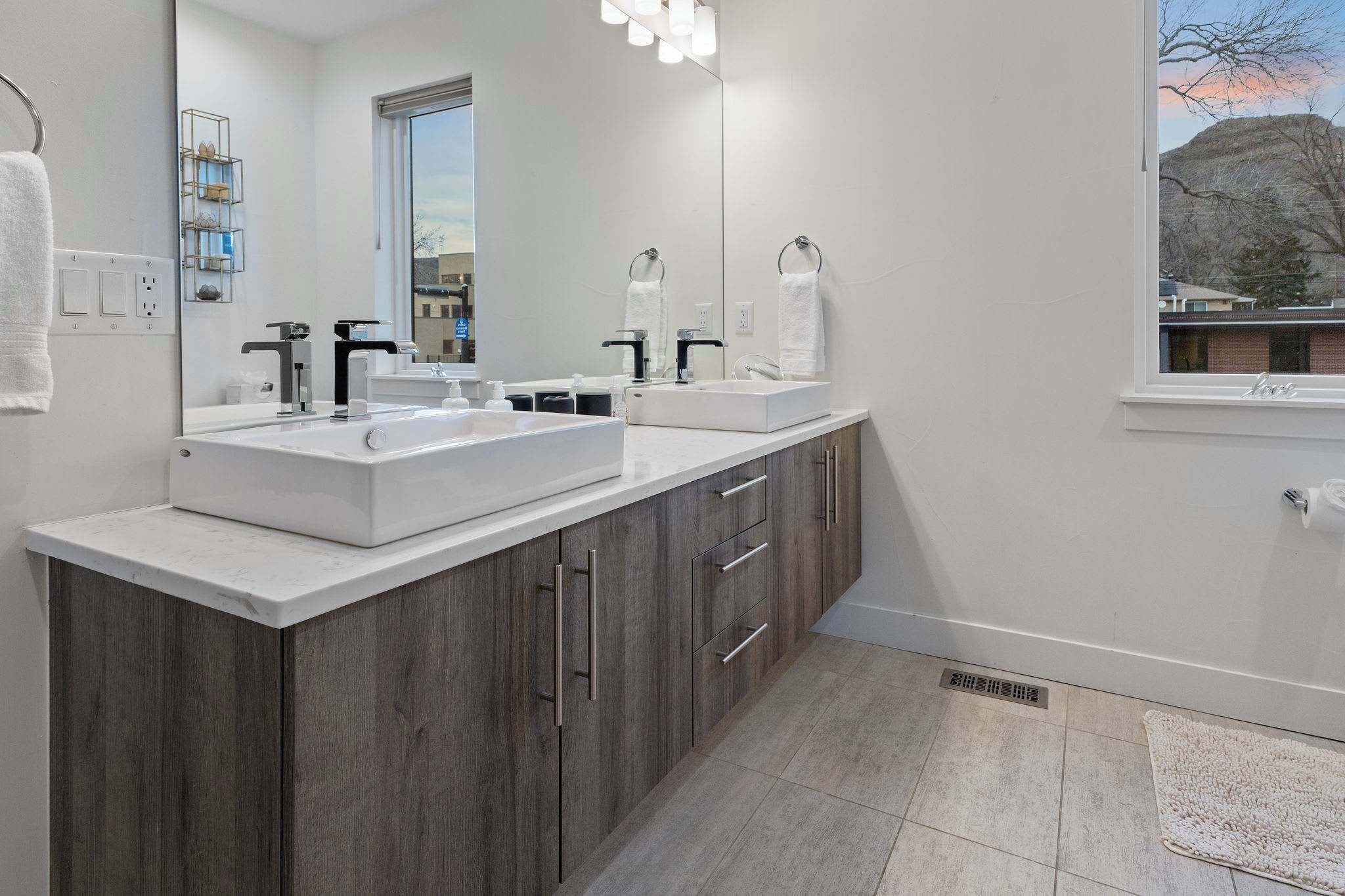 A modern bathroom with a large mirror, two sinks, wooden cabinets, and a window with a view of a building and trees.