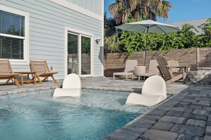 Une piscine entourée d’une clôture en bois et de verdure, avec des chaises longues et un parasol.