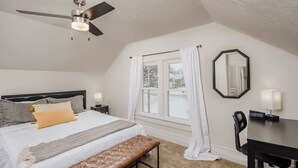Een slaapkamer met een bed, een bankje, een bureau, een stoel, een plafondventilator, een raam met gordijnen en een spiegel.