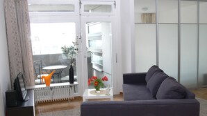 Uma sala de estar com um sofá cinzento, uma mesinha com um vaso de flores, um radiador e vista para uma varanda com cadeira e mesa.