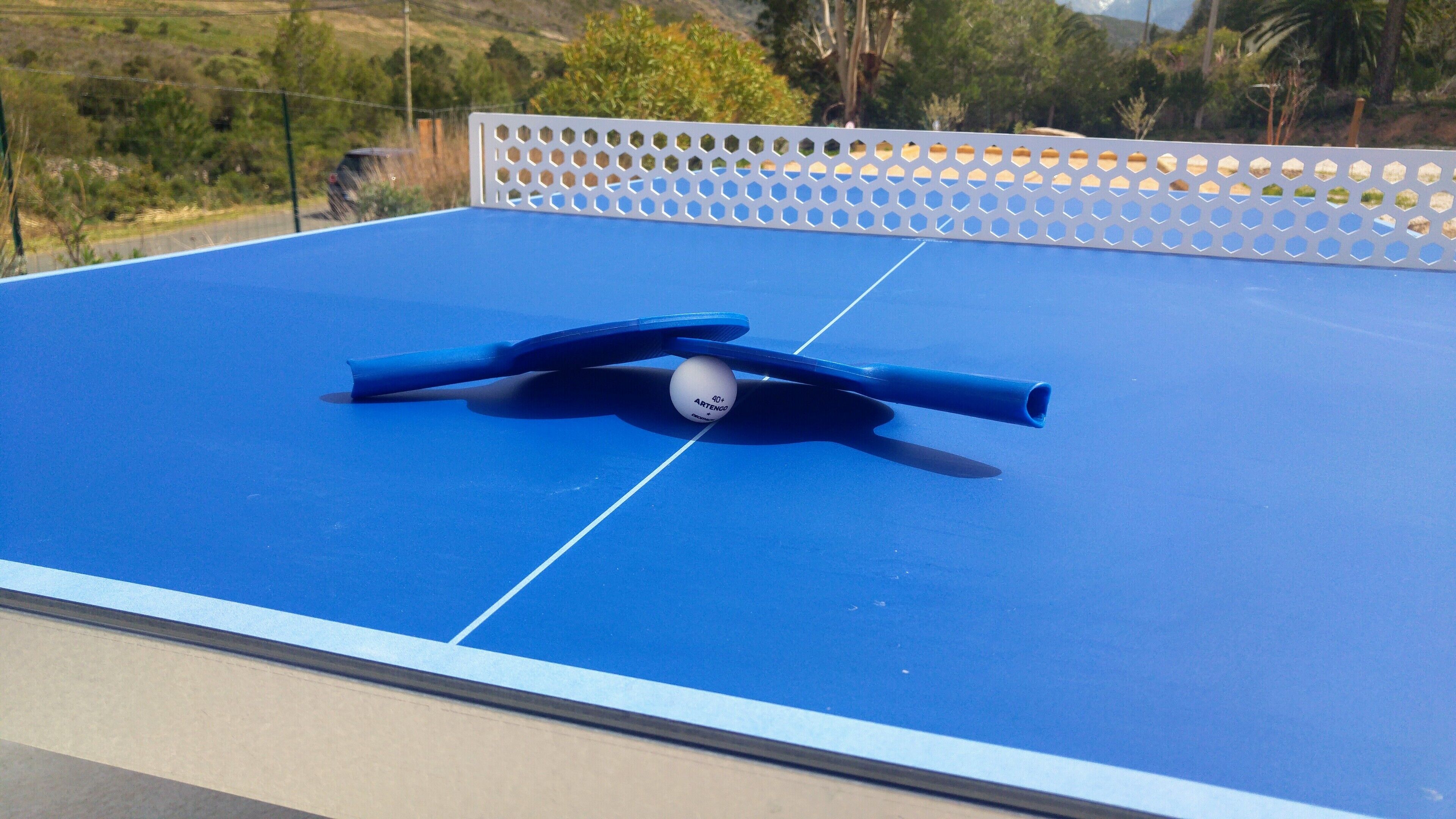 A blue ping pong table with a ball and paddles, surrounded by a white fence.