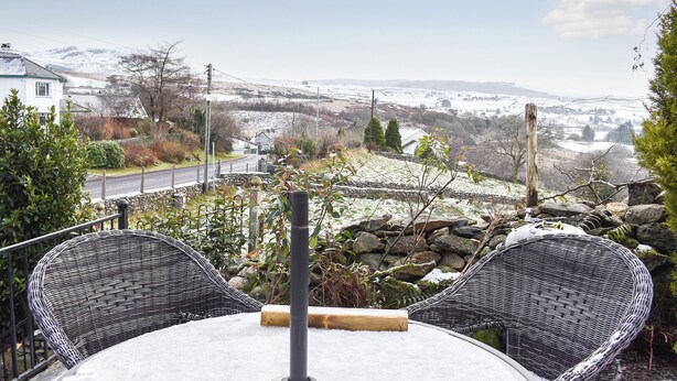 Un patio con dos sillas de mimbre y una mesa, con vistas a un paisaje nevado que incluye una carretera y casas.