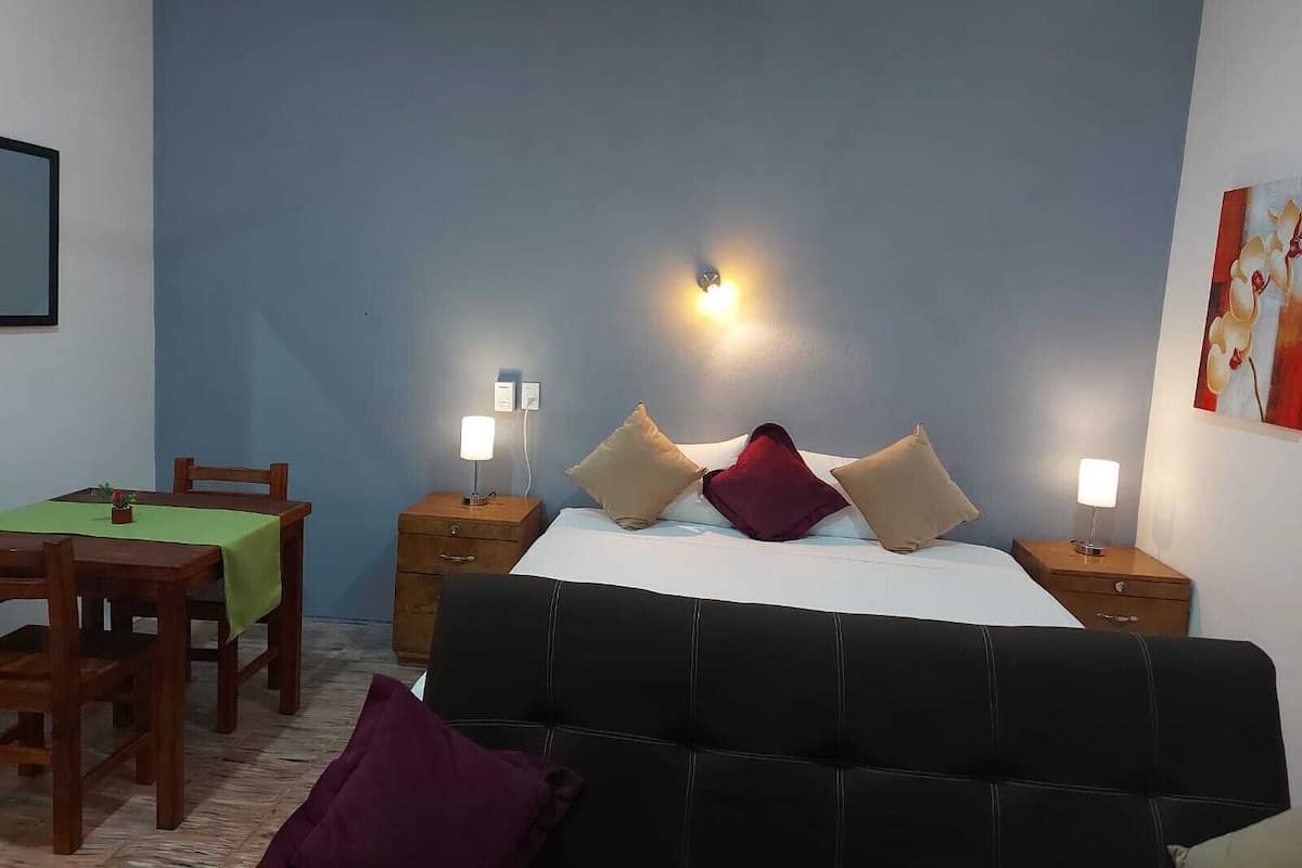 Un dormitorio con cama, dos mesitas de noche con lámparas, una mesita auxiliar de madera, una silla y una mesa con mantel verde.