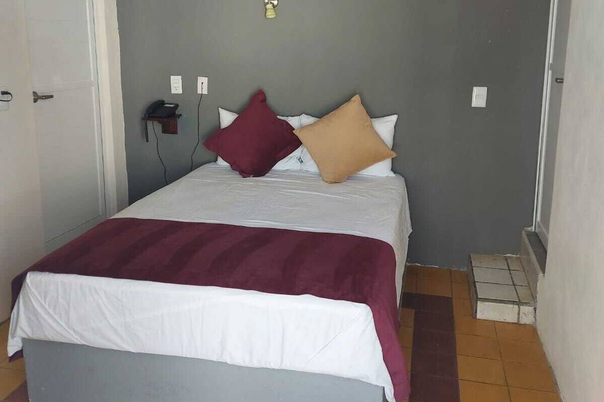 Una cama individual con una colcha blanca y granate, dos almohadas, un teléfono en la mesita de noche y una lámpara de pared.