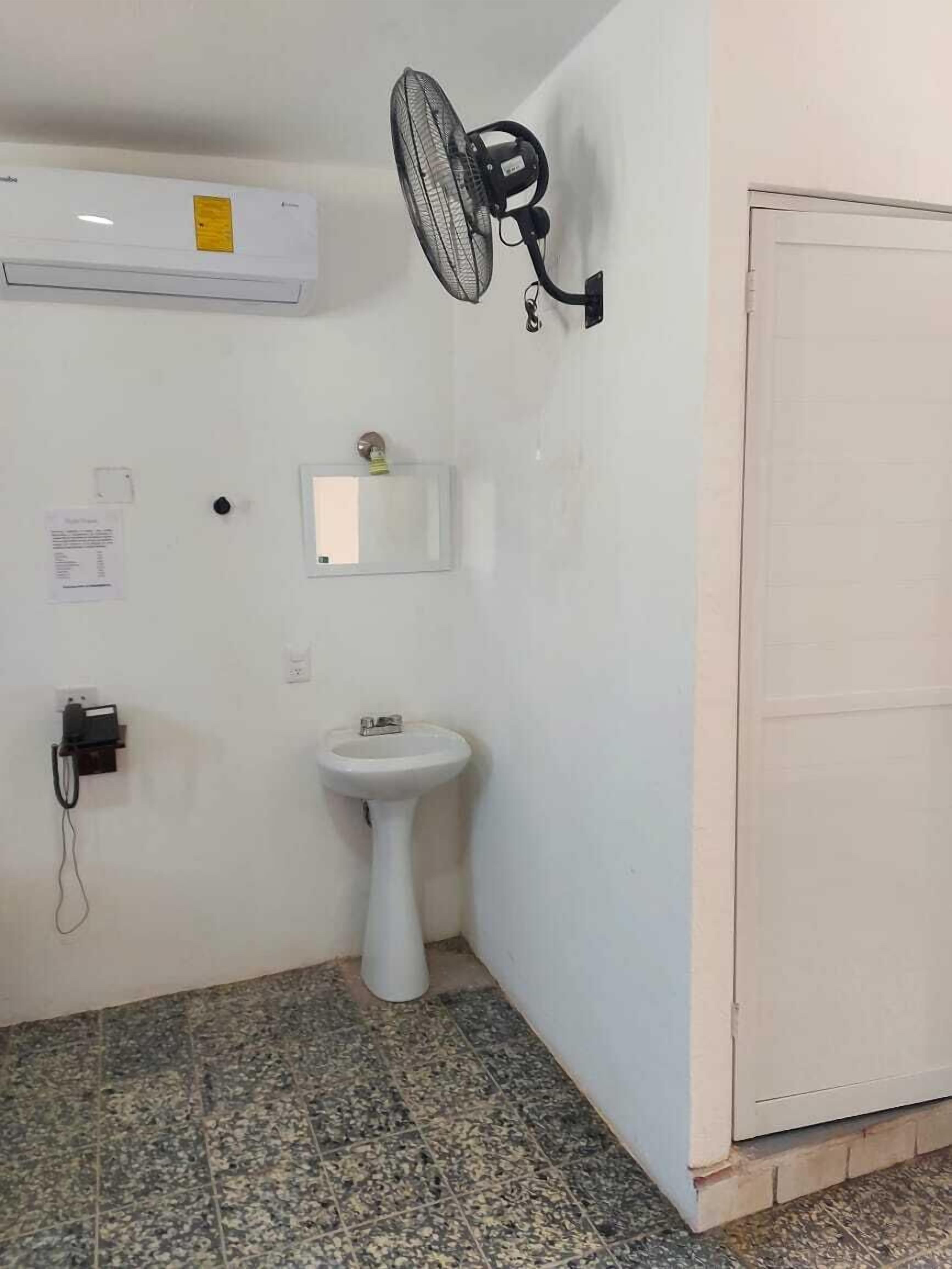 Un baño pequeño con un lavamanos pedestal, un ventilador de pared y una puerta blanca.