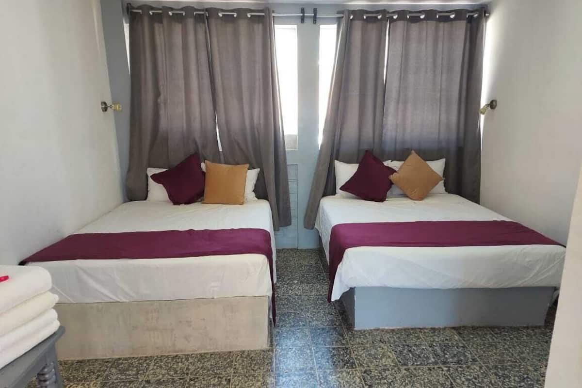 Habitación con dos camas, cada una con colchón blanco y manta color burdeos. El piso es de baldosas y las cortinas son grises.