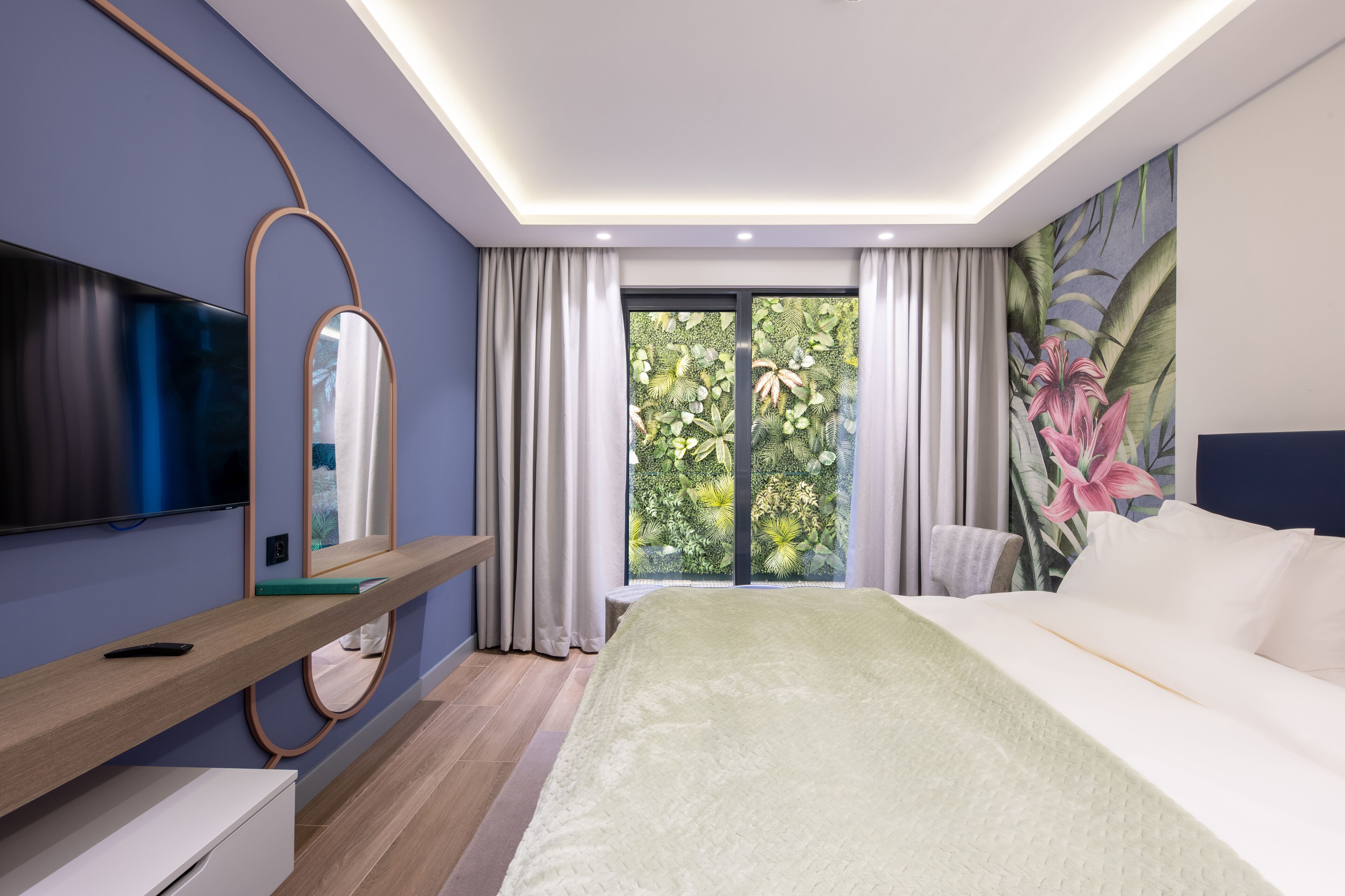 A modern hotel room with a large bed, a flat-screen TV, a wooden bedside table, a mirror, and a view of lush greenery through the window.