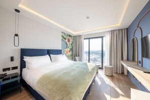 A modern hotel room with a large bed, a flat-screen TV, a desk, and a balcony with a view.