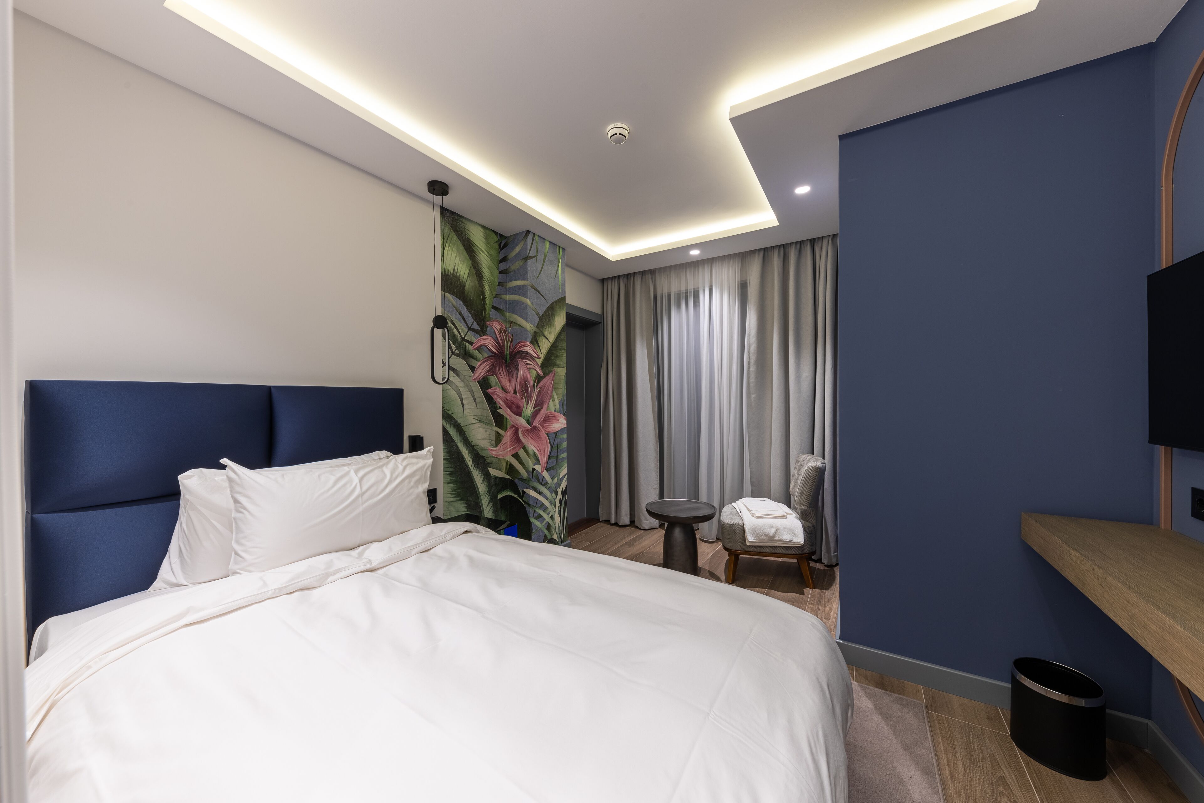 Una habitación de hotel moderna con una cama grande, un cabecero, un televisor montado en la pared, una mesita, una silla y una cortina.
