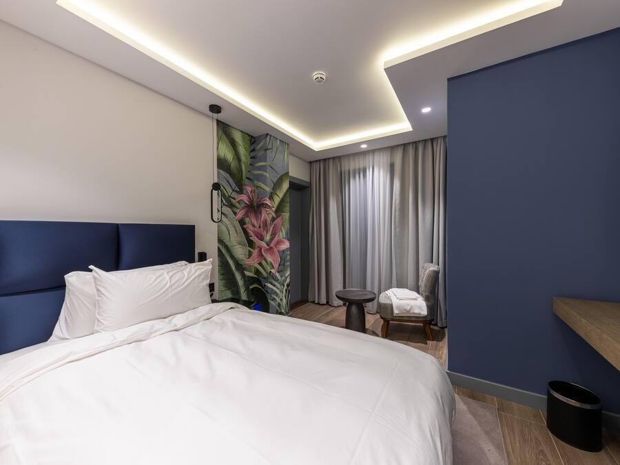 A modern hotel room with a large bed, a headboard, a wall-mounted TV, a small table, a chair, and a curtain.