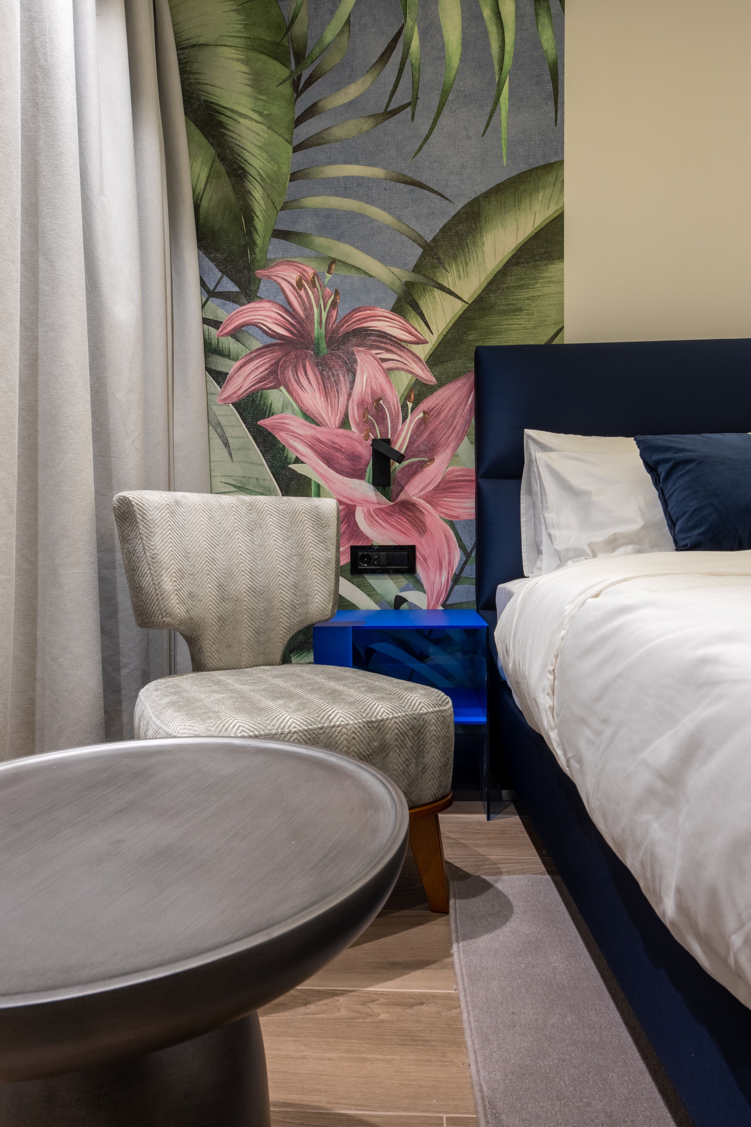 A bedroom with a bed, a chair, a small table, and a wall with a floral mural.