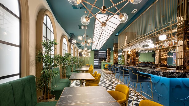 Un restaurant spacieux avec de hauts plafonds, des sièges recouverts d’un tissu vert et un bar agrémenté de touches de bleu.